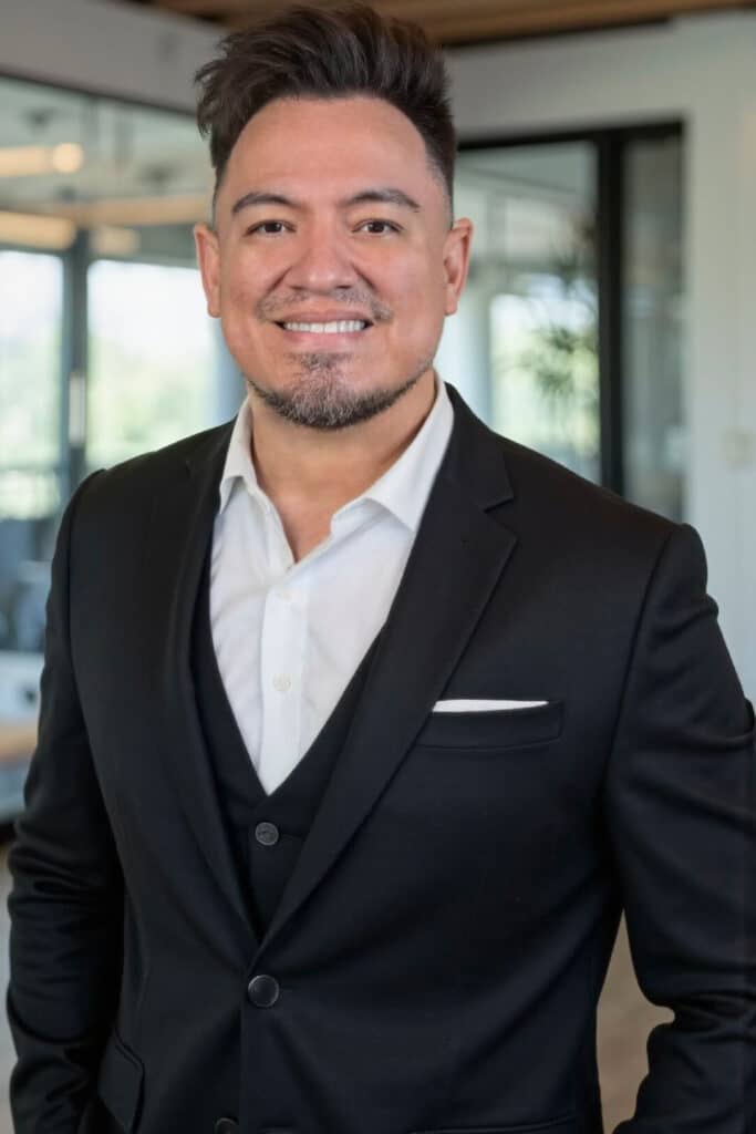 Carlos-Garcia-Photo | Search Quest A man with short dark hair and a trimmed beard smiles while wearing a black suit with a white shirt and pocket square, standing in a modern office setting with large windows and glass walls.