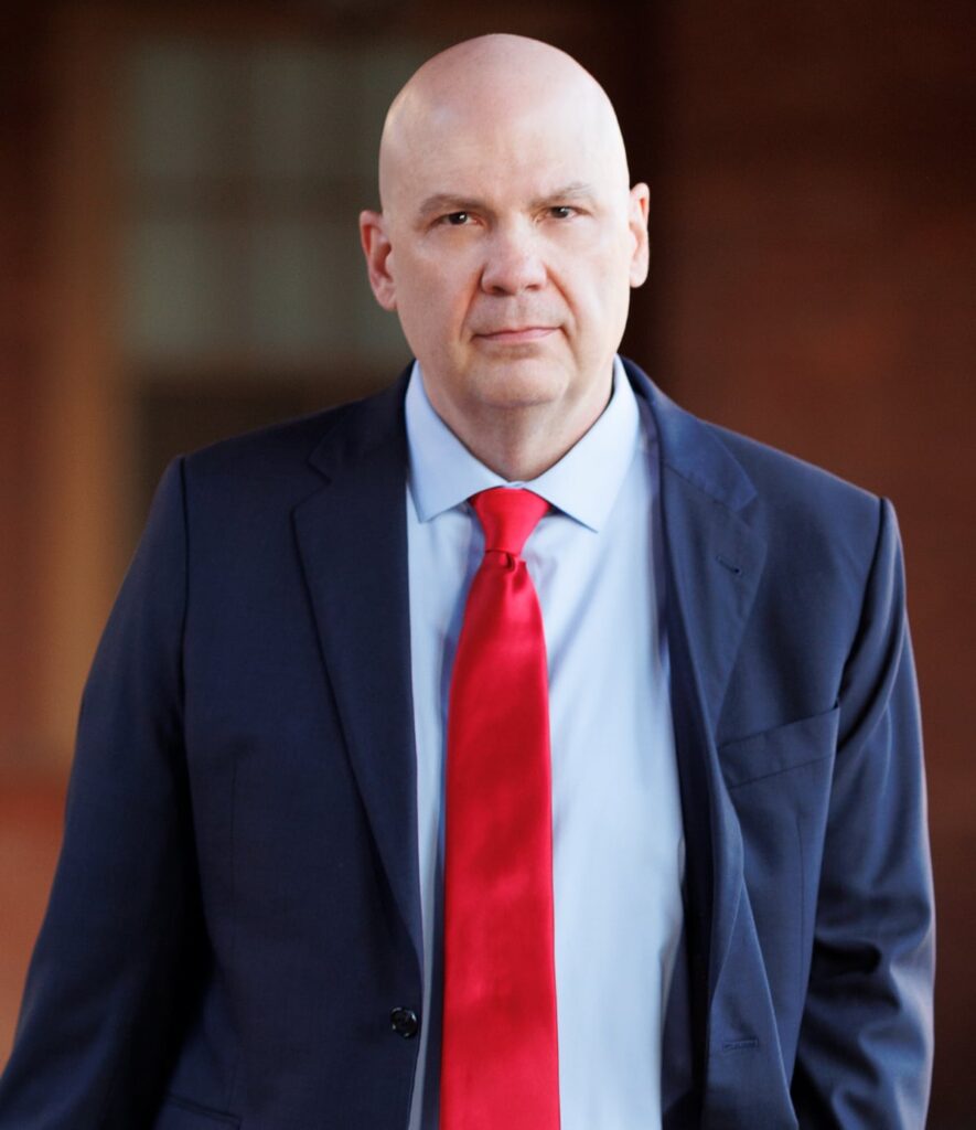 roofing-gary | Search Quest A bald man wearing a dark blue suit, light blue shirt, and bright red tie stands outdoors, looking directly at the camera with a neutral expression.