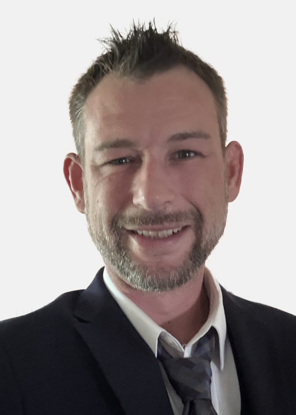 A man with short hair and a trimmed beard wearing a dark navy suit jacket, white shirt, and patterned grey tie, smiling at the camera against a plain light background.